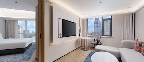 A hotel room with a bed, a sofa, a TV mounted on the wall, and a view of the cityscape through the windows.