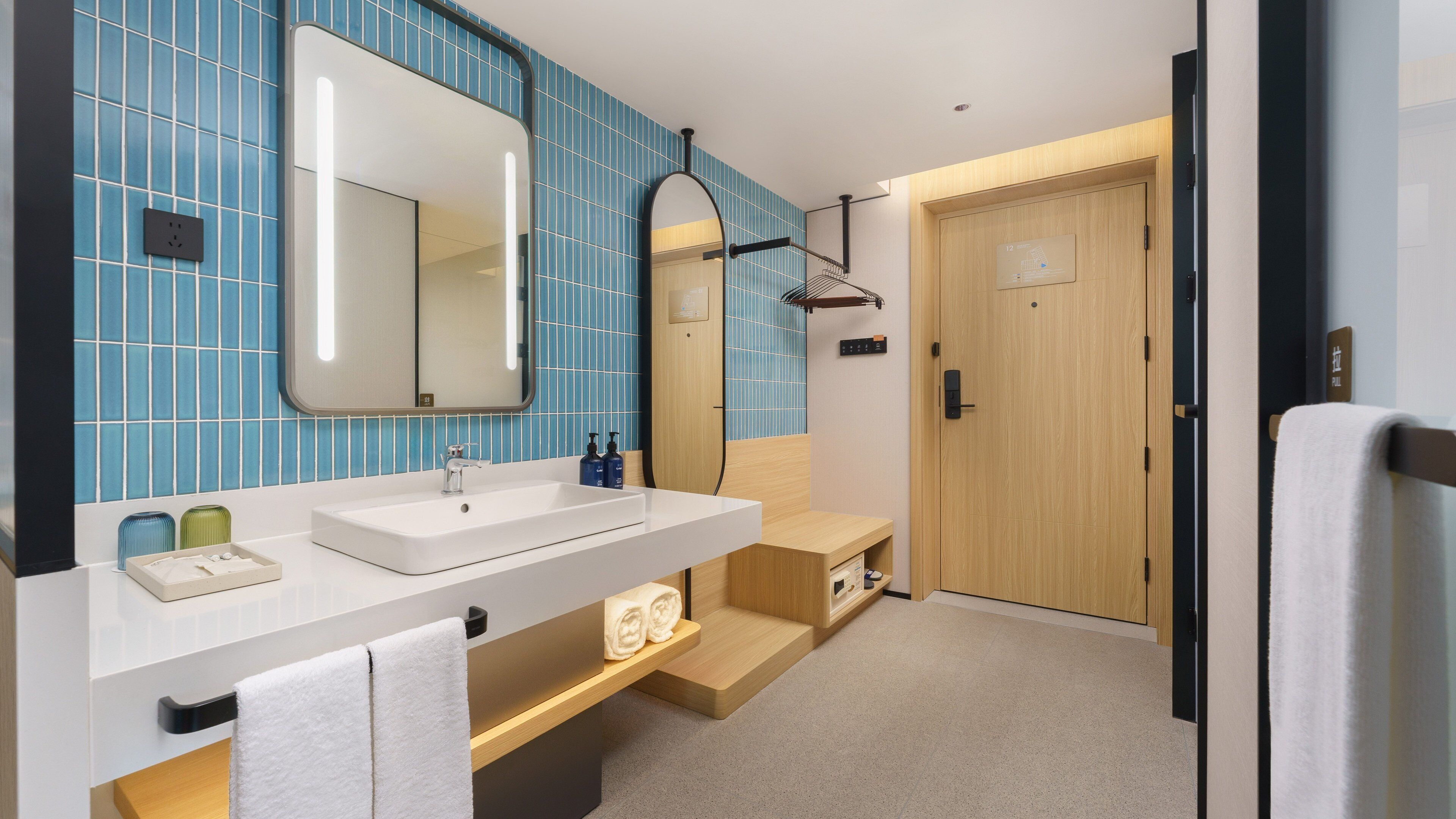 A modern bathroom with a white sink, blue-tiled walls, a large mirror, and a wooden door.