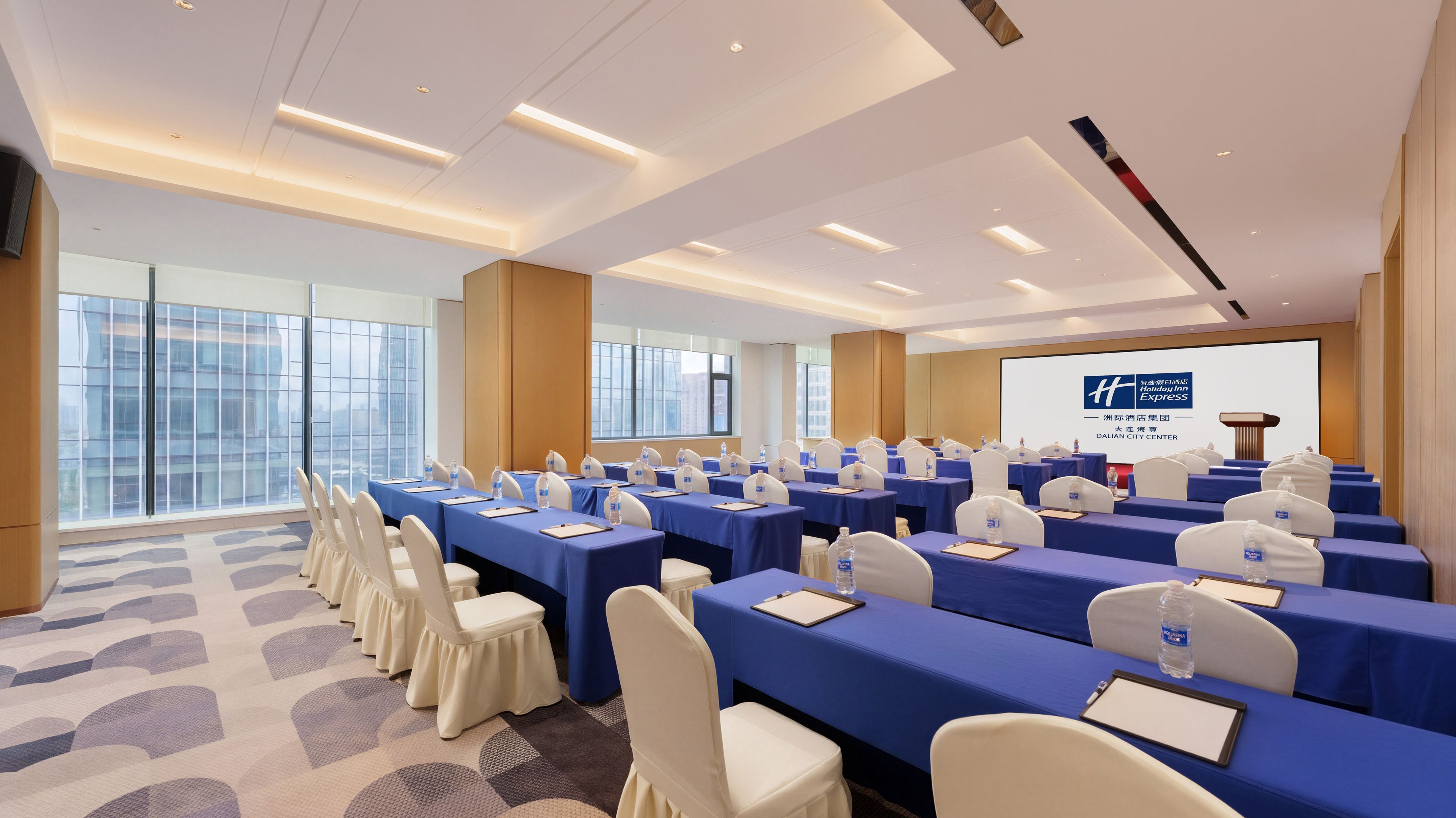 A conference room with rows of tables and chairs, a projector screen, and a view of the city through large windows.
