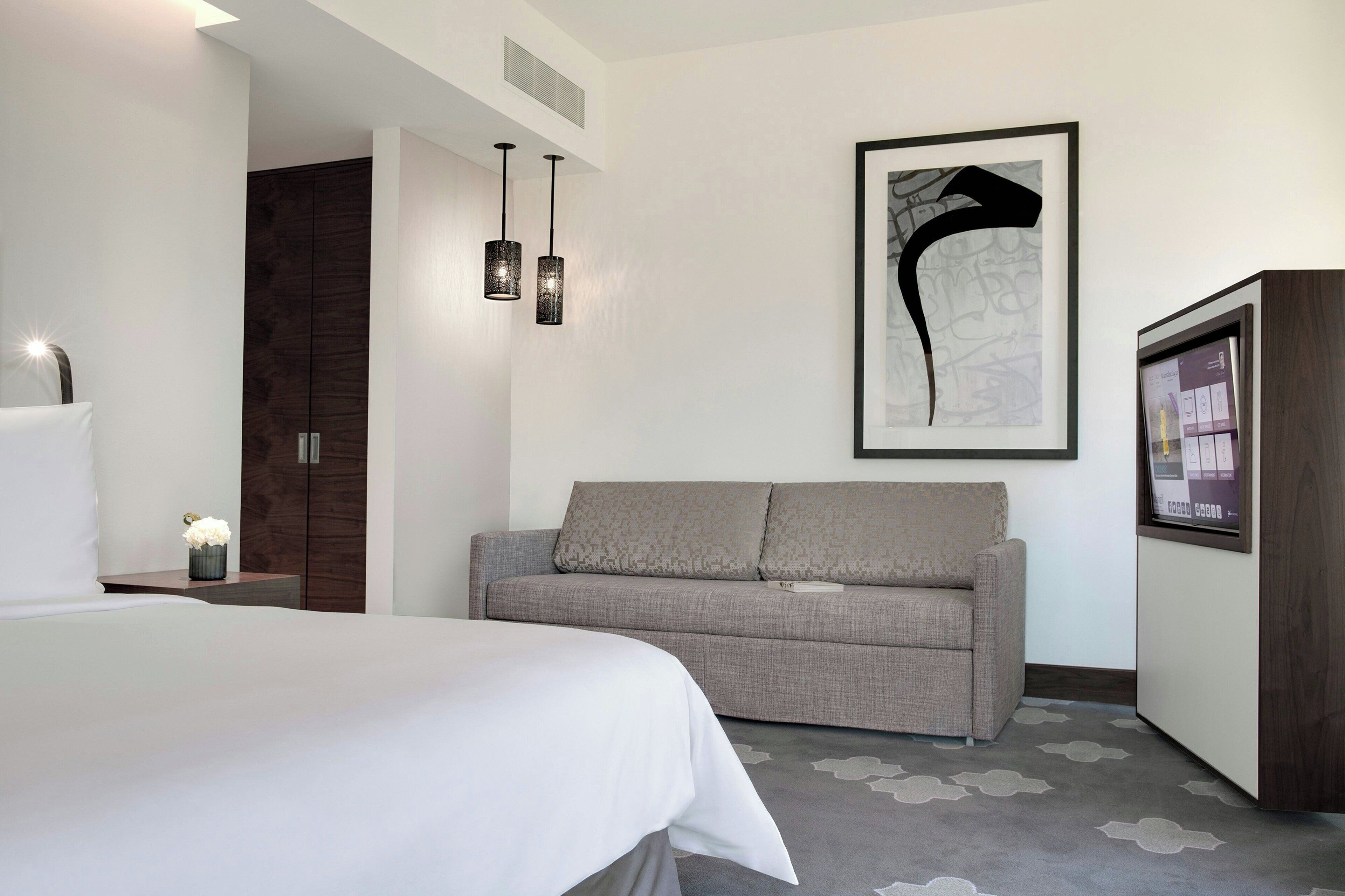 Une chambre d’hôtel moderne avec un grand lit, un canapé gris, une télévision et une œuvre d’art abstraite encadrée accrochée au mur.