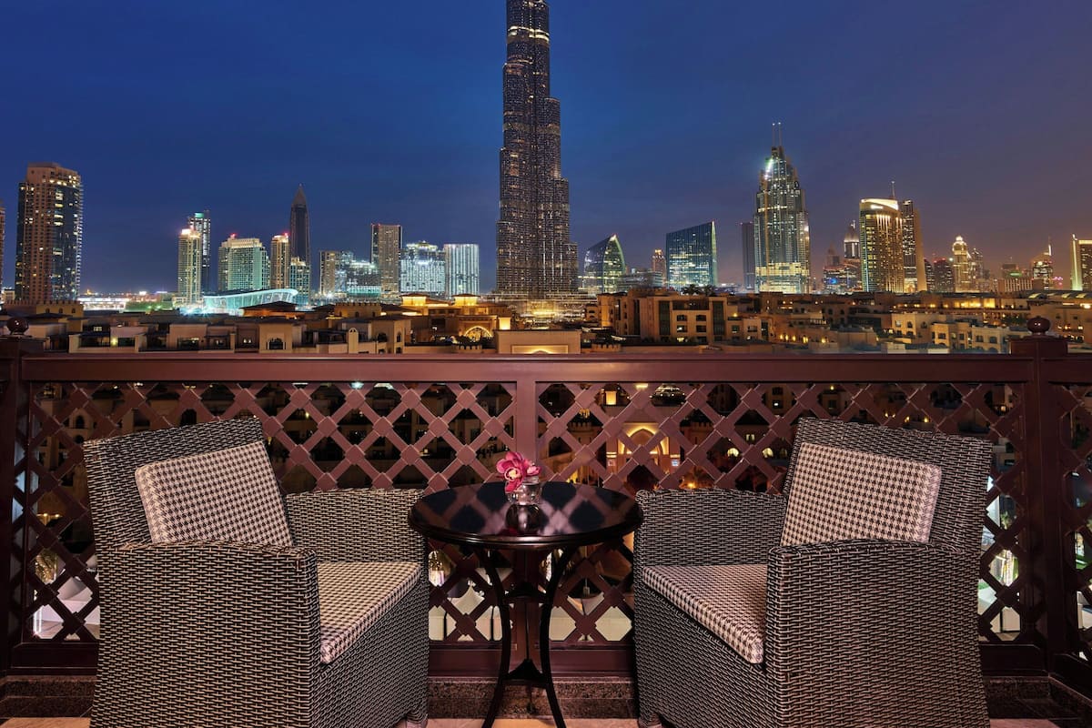 A balcony with wicker furniture and a table, overlooking a city skyline at night.