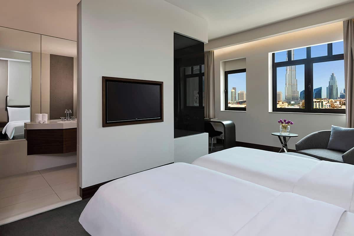A hotel room with a bed, a TV, a desk, a chair, a small table with a vase, and a window with a city view.