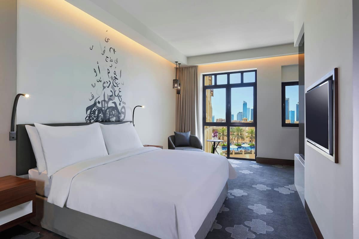A modern hotel room with a large bed, a desk, a chair, a TV, and a view of the city through large windows.