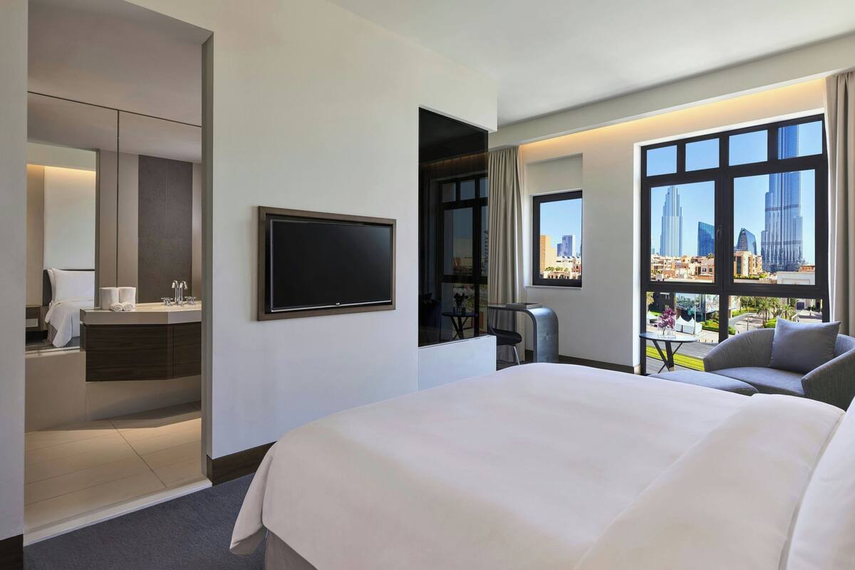 A hotel room with a large bed, a TV, a desk with a chair, a bathroom, and a city view.