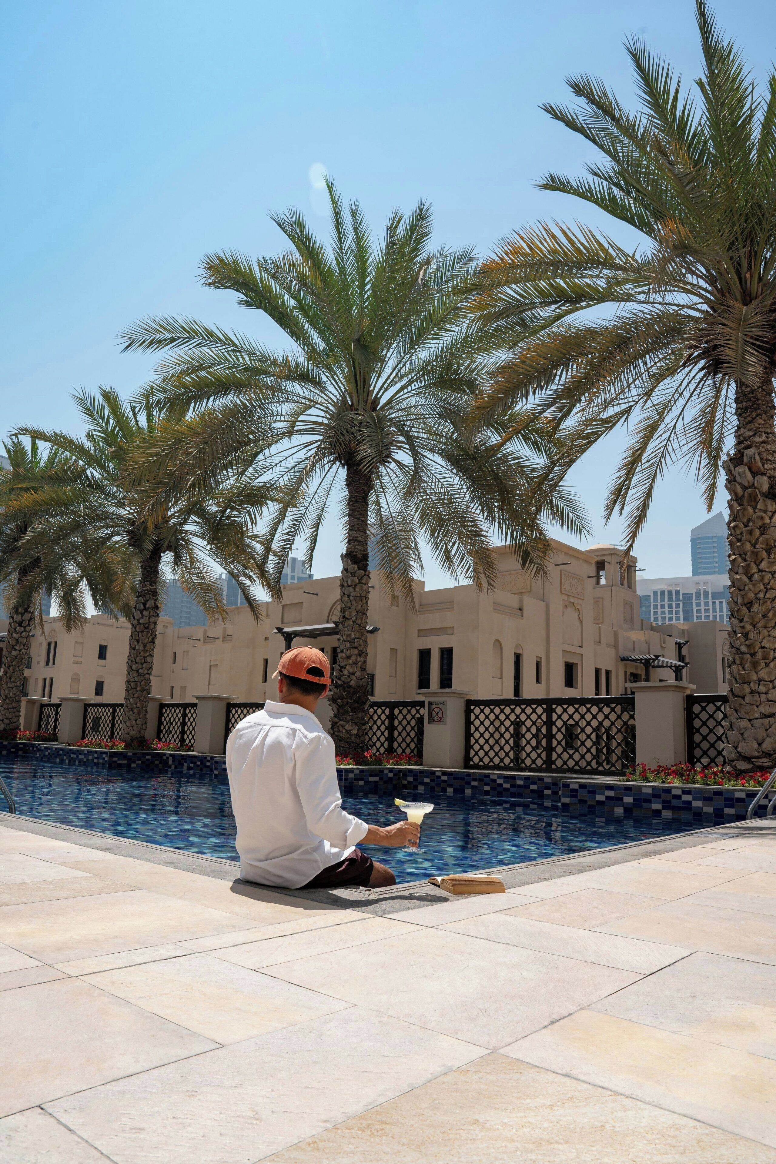 A man sitting by a pool with palm trees and a building in the background.