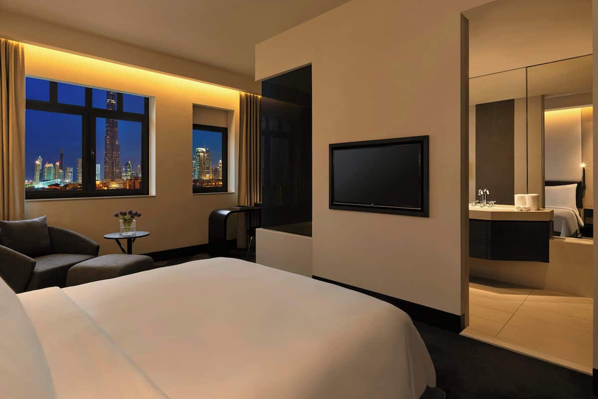 A hotel room with a large bed, a TV, a seating area with a table and chairs, and a view of a city skyline at night.