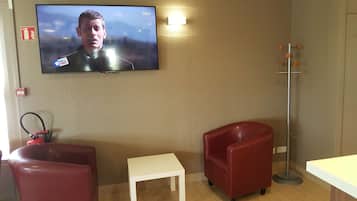 Een kamer met een aan de muur bevestigde televisie, een klein wit tafeltje, twee rode stoelen en een vloerlamp.