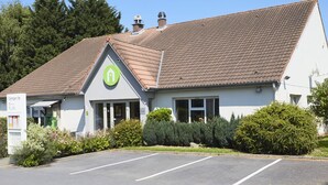 A single-story building with a sloped roof, a green logo on the front, and a parking lot in front. - Campanile Montluçon - St Victor (Saint-Victor)