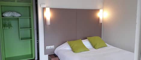 Una cama con ropa de cama blanca y dos almohadas verdes, una mesita de noche, un armario verde con prendas colgadas y dos luces fijadas a la pared.