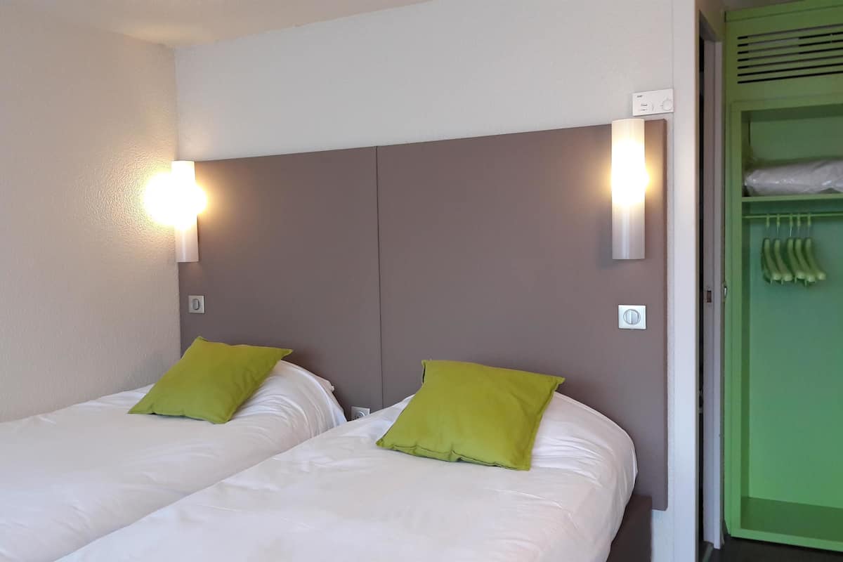 Ein Hotelzimmer mit zwei Einzelbetten, jeweils mit einem grünen Kopfkissen und einer grauen Kopfteil, sowie einer wandmontierten Lampe.