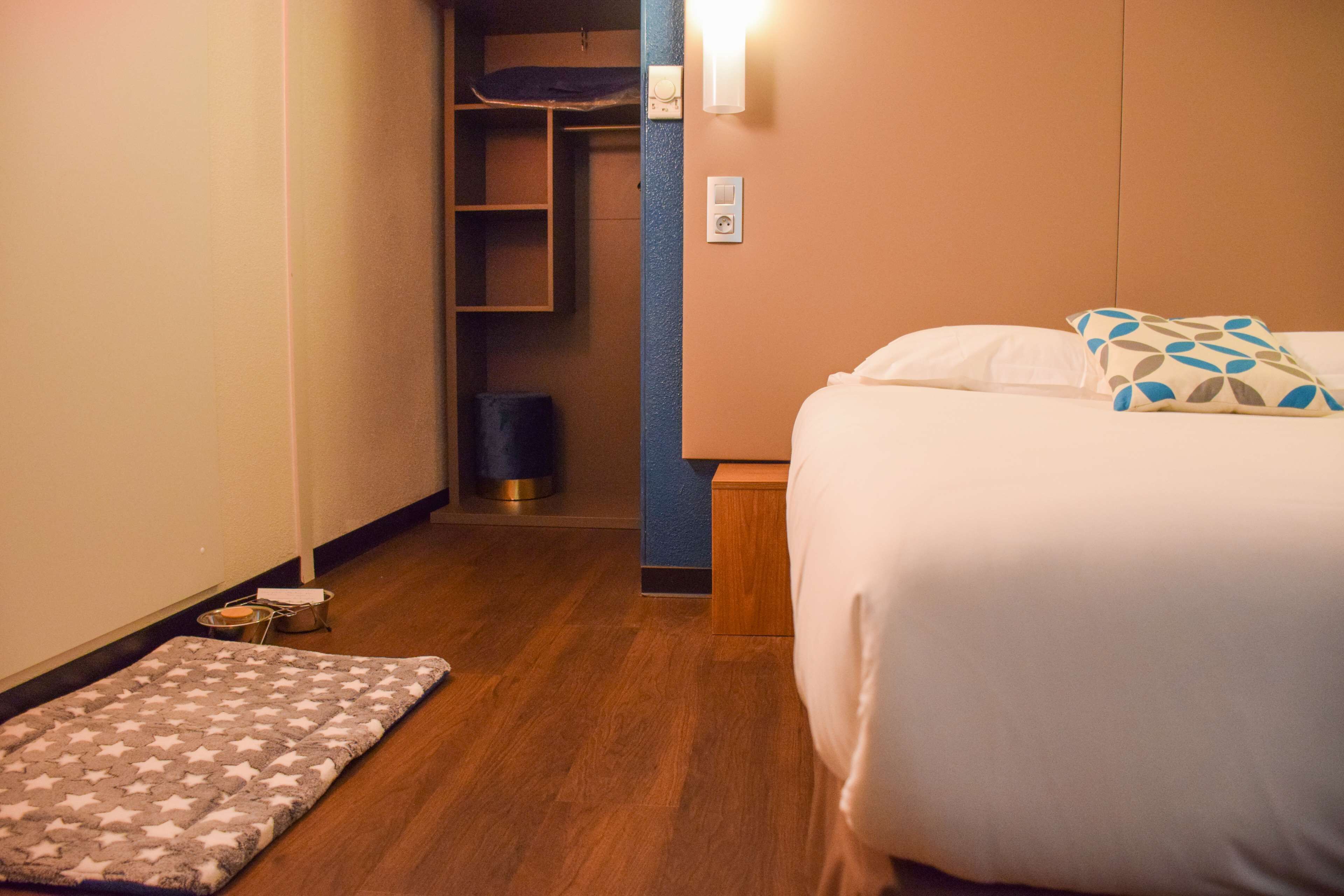Ein Hotelzimmer mit Bett, Nachttisch, Teppich, Kleiderschrank und Mülleimer.