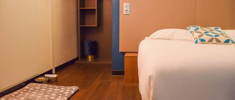A hotel room with a bed, a bedside table, a nightstand, a small rug, a closet, and a trash can.