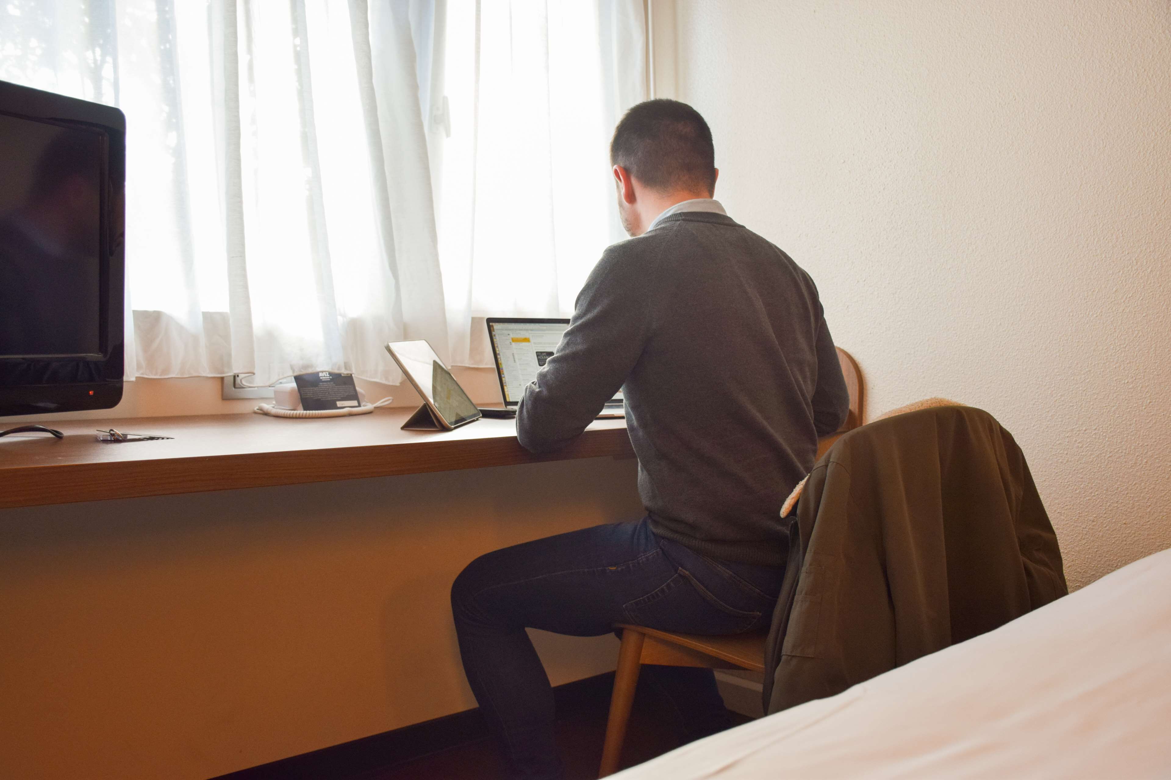 A man sitting at a desk with a laptop, a smartphone, and a tablet. There is a TV on the left and a bed in the background.
