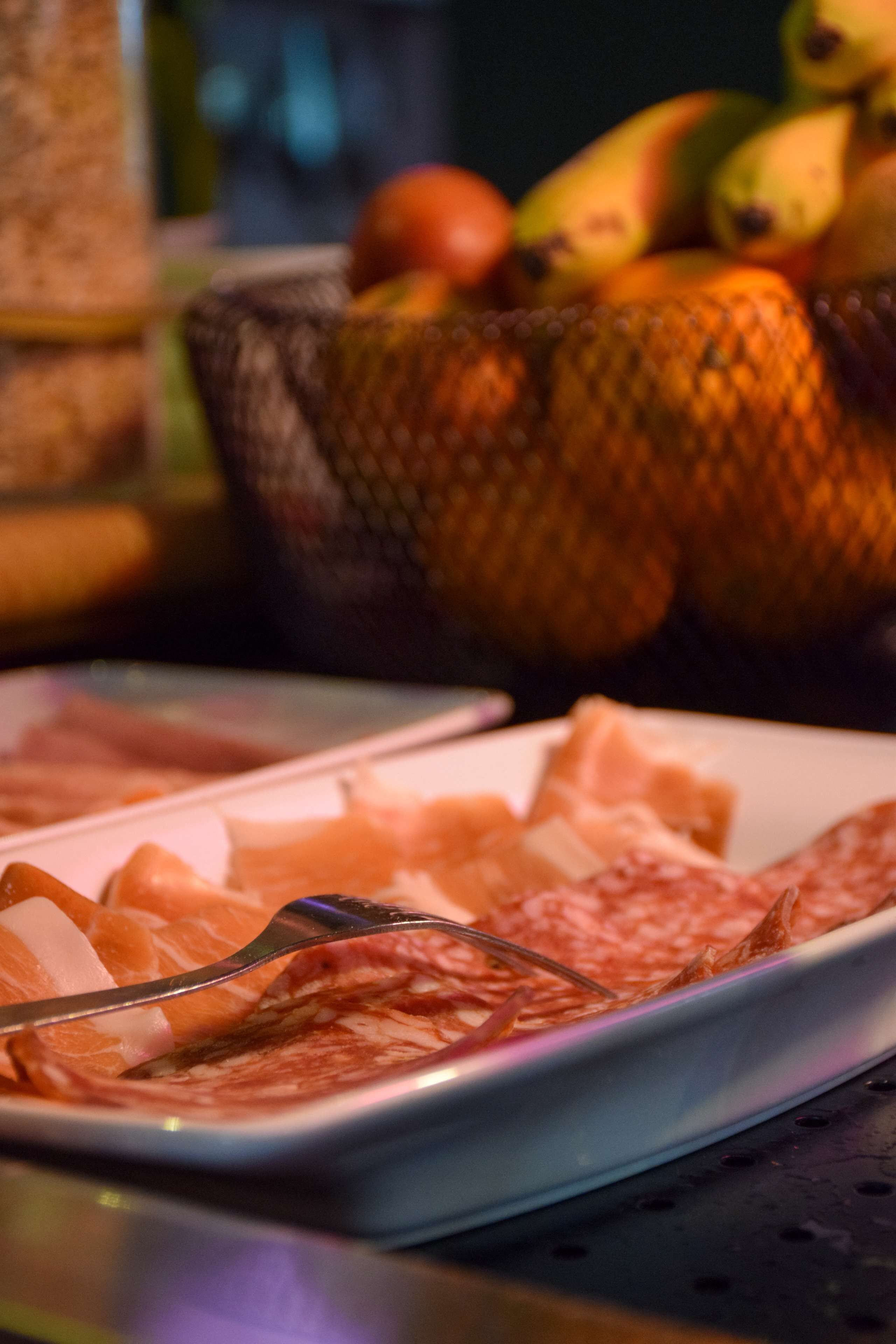 Une assiette de charcuteries et un panier de fruits sont posés à l’arrière-plan.