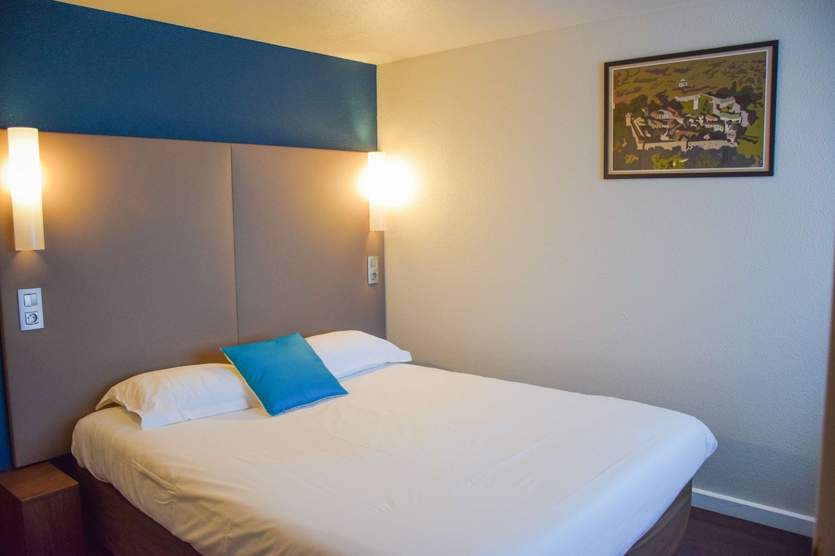 A neatly made bed with white bedding and a blue pillow, two wall-mounted light fixtures, and a framed picture on the wall.