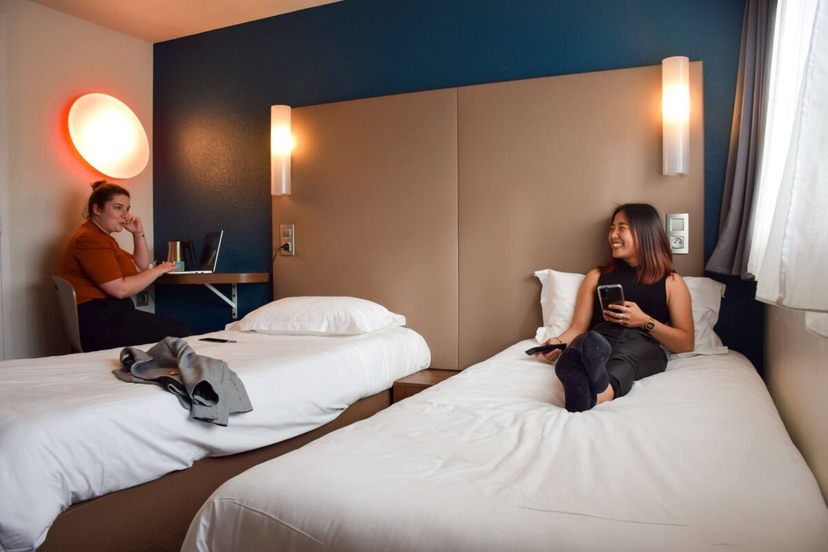 Zwei Einzelbetten in einem Hotelzimmer mit Schreibtisch, Stuhl, einer Wandlampe und einem mit Vorhängen versehenen Fenster.