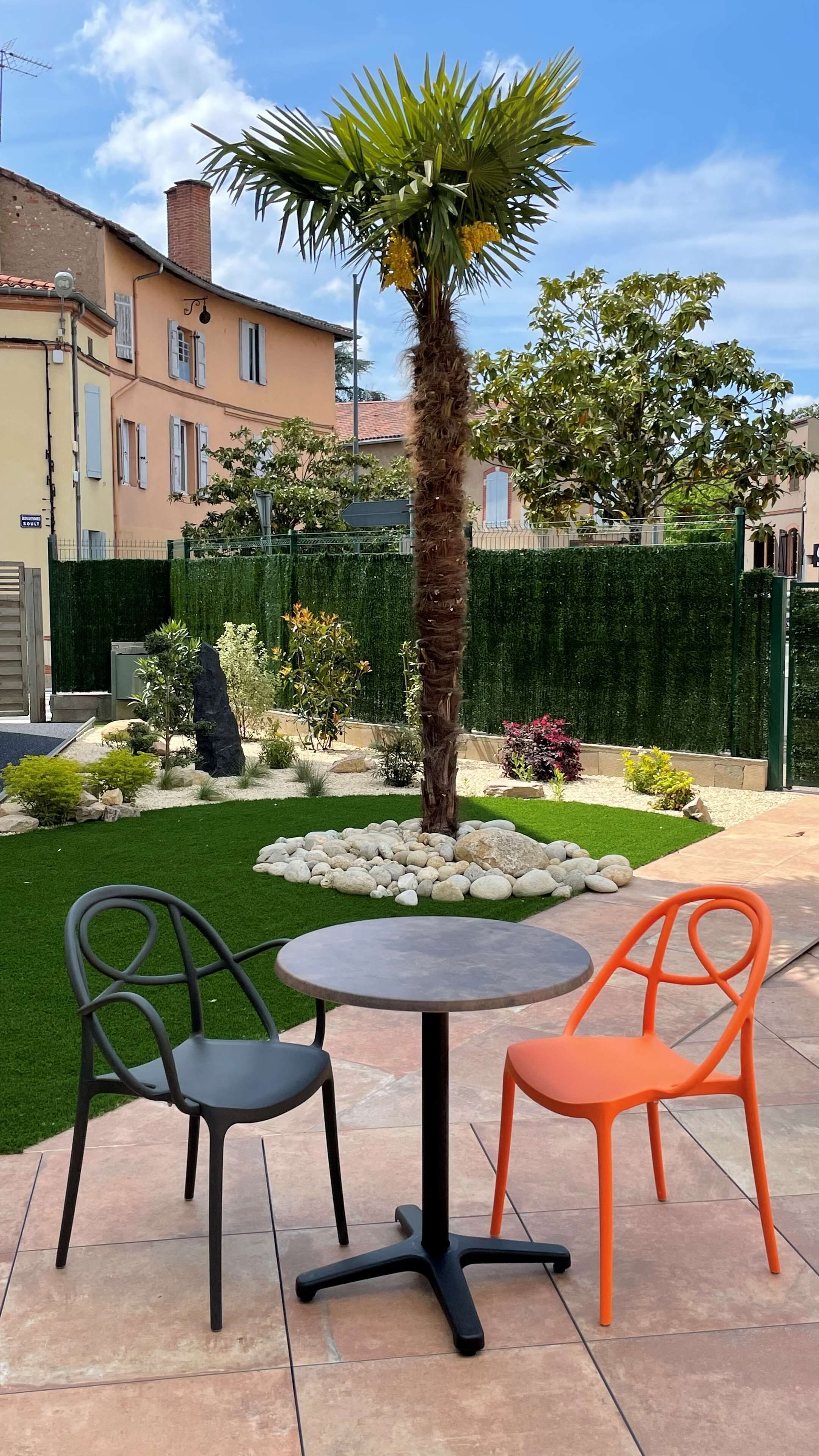 An outdoor seating area with two chairs and a table, surrounded by a well-maintained garden with a palm tree, various shrubs, and a hedge.