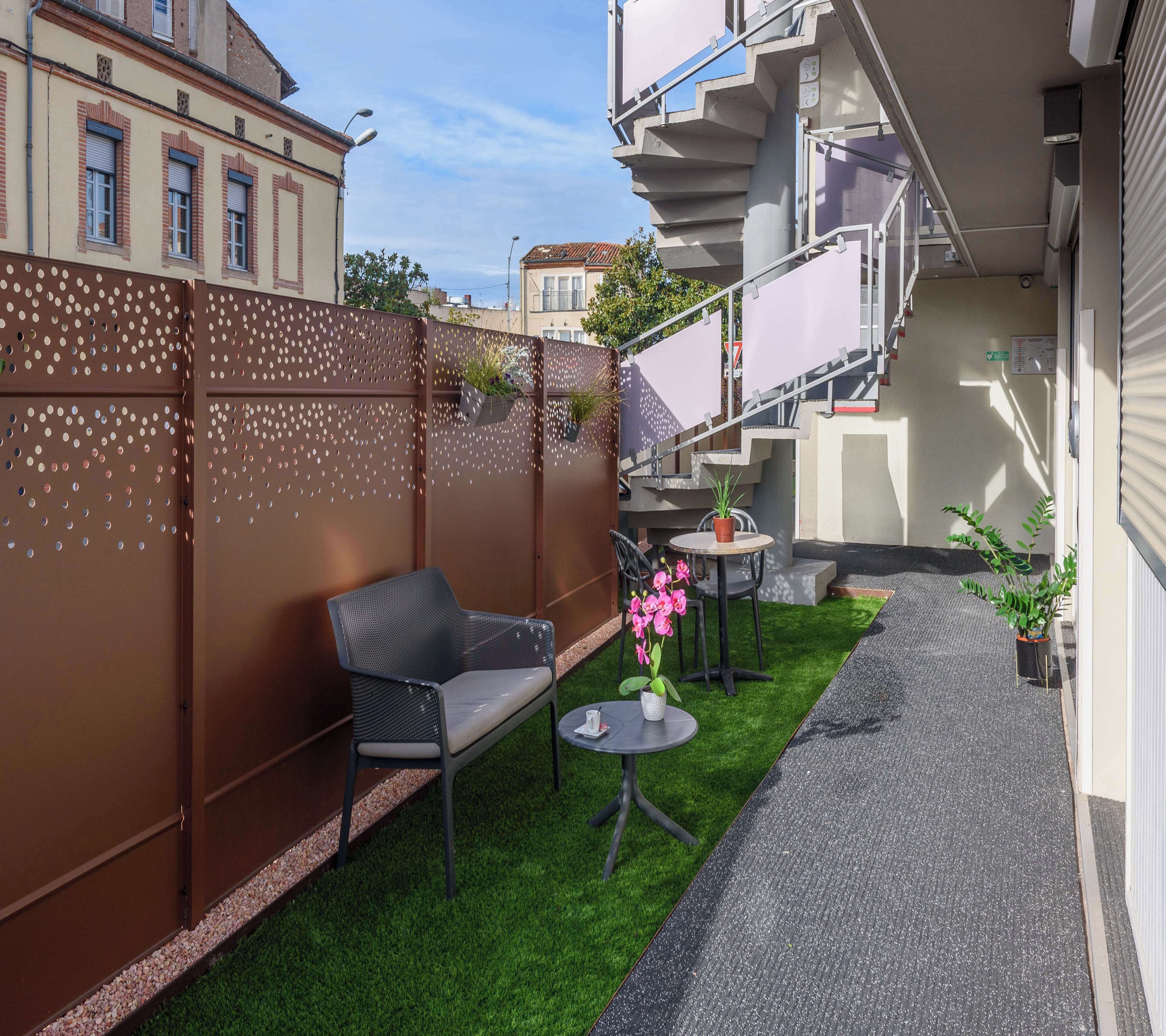Zona de pati exterior petita amb gespa artificial, una cadira de metall negre, una taula rodona petita i una planta en test amb flors roses.