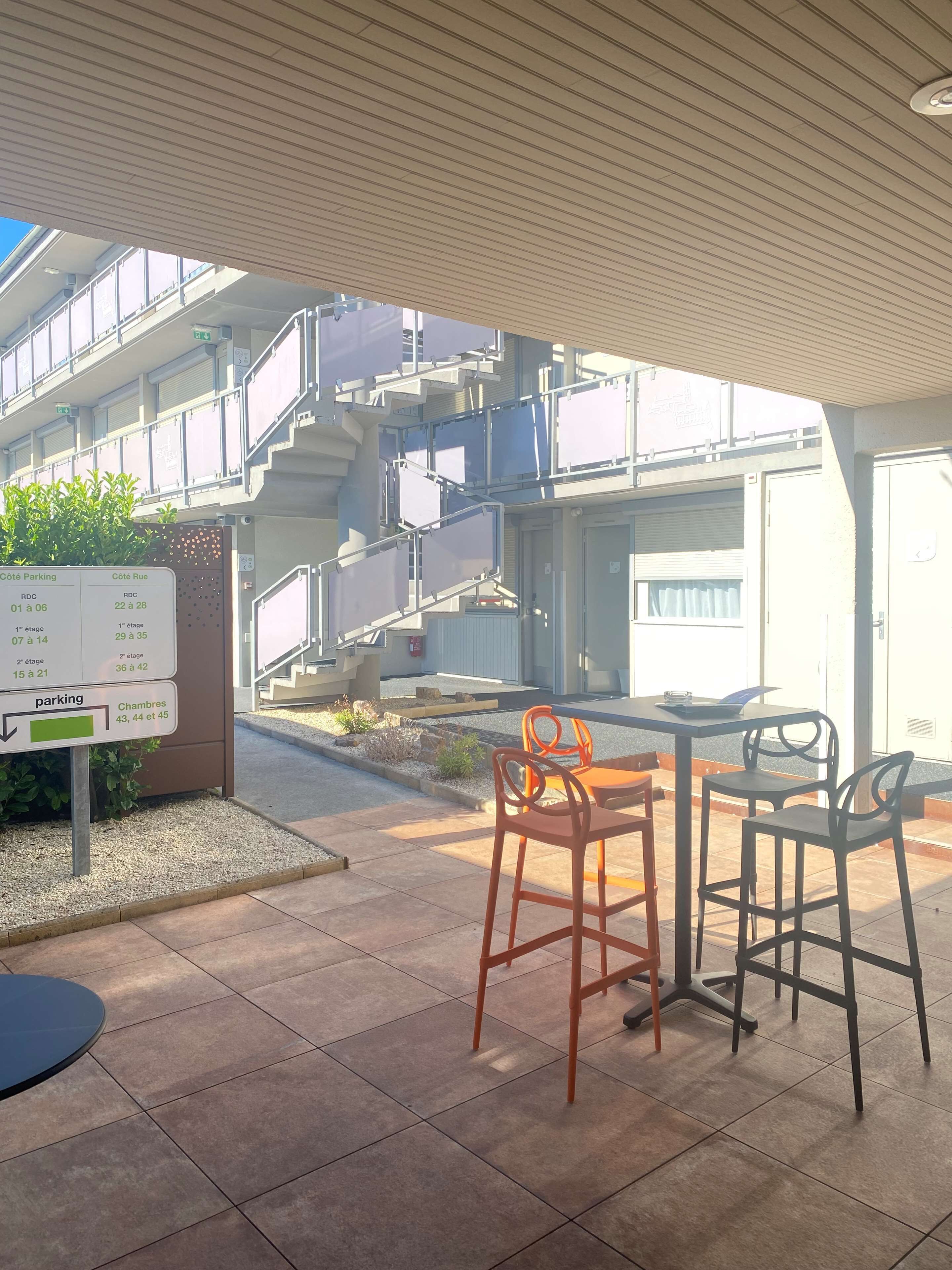 Een buitenzitgedeelte met een tafel en stoelen, een bord dat de parkeerlagen aangeeft, en een gebouw met meerdere verdiepingen en balkons op de achtergrond.