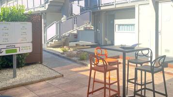 Een buitenzitgedeelte met een tafel en stoelen, een bord dat de parkeerlagen aangeeft, en een gebouw met meerdere verdiepingen en balkons op de achtergrond.