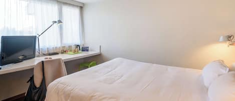 A hotel room with a bed, a desk with a computer, a lamp, and a window with curtains.