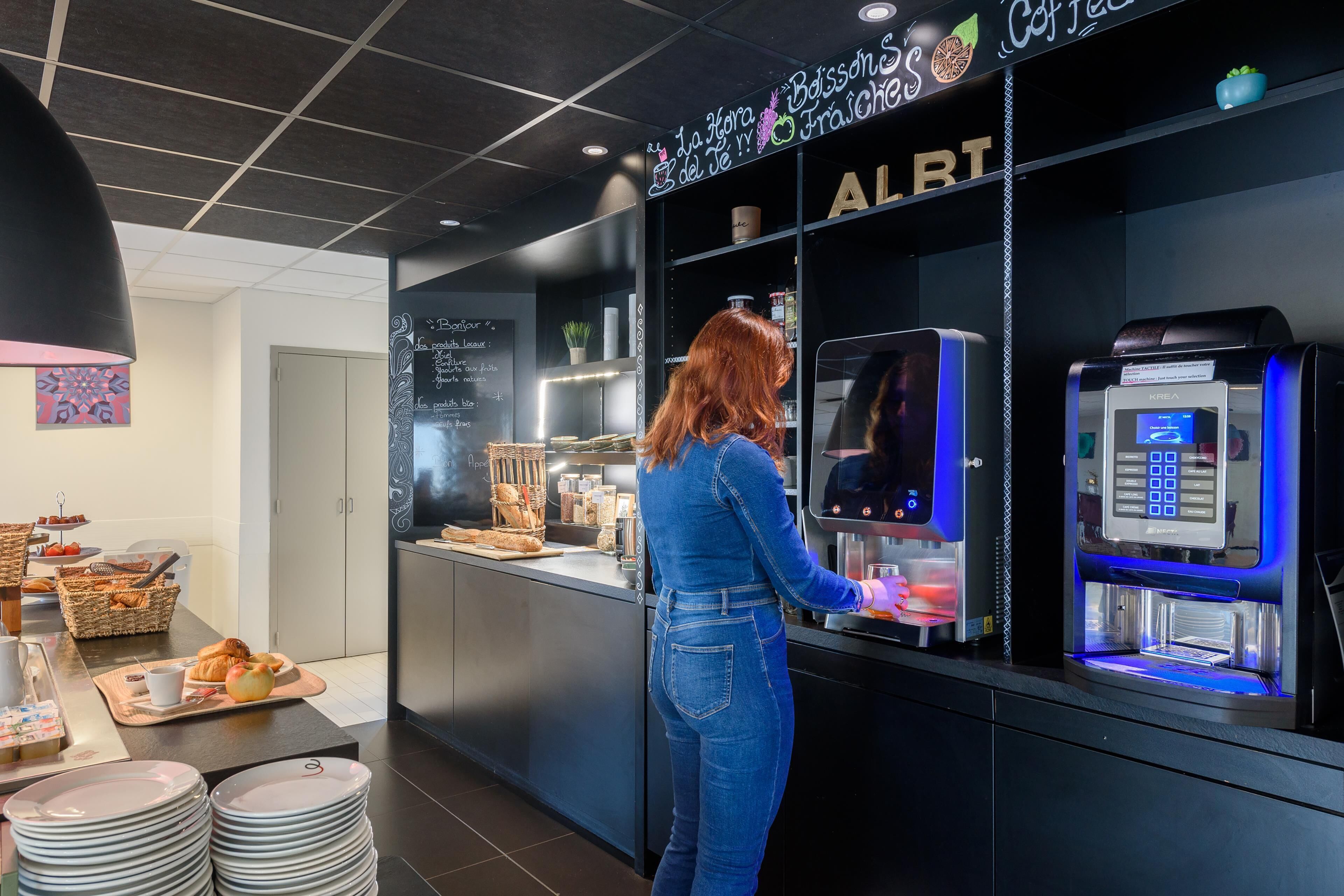 A woman using a coffee machine in a modern cafe setting with a counter displaying pastries and a chalkboard menu.