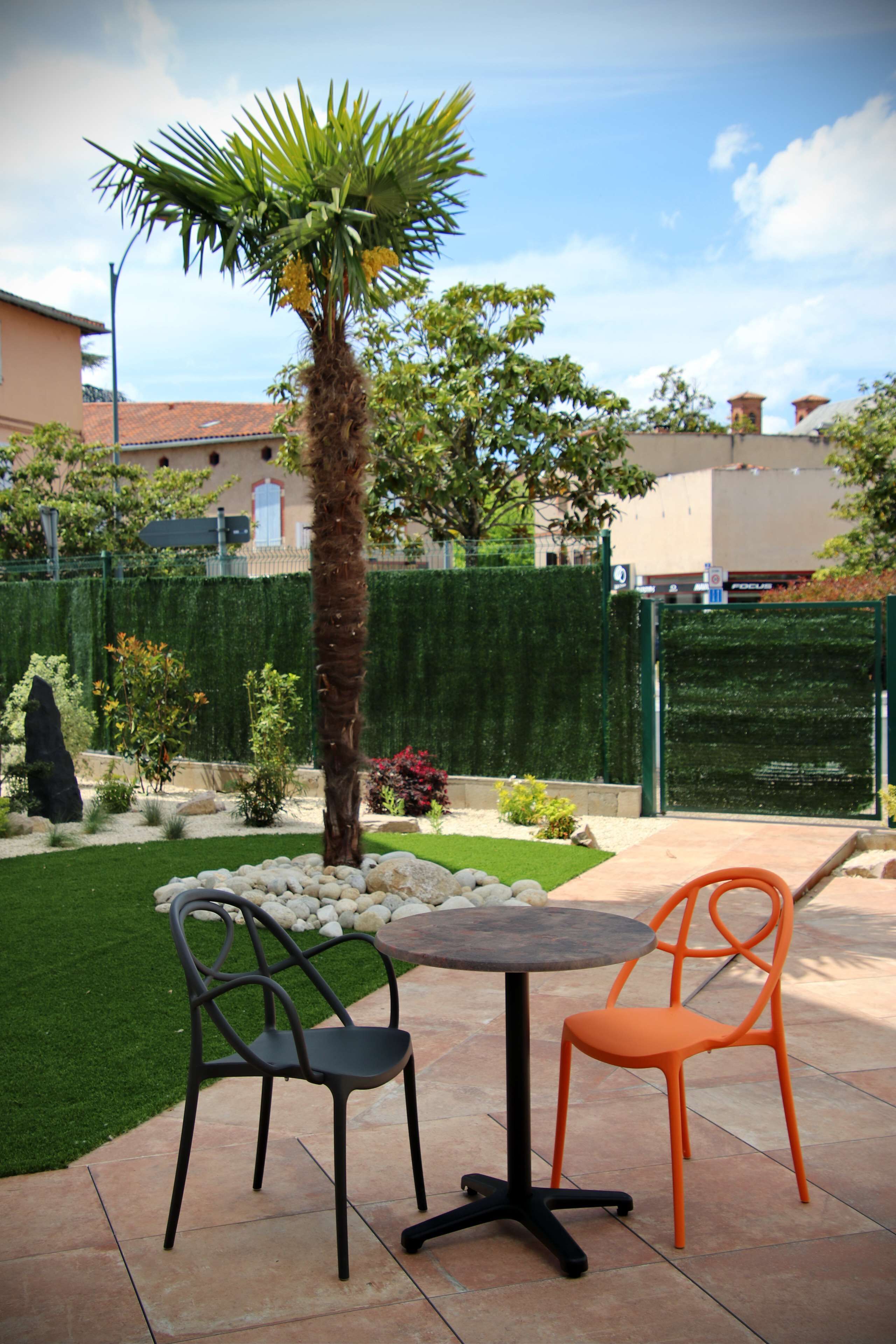 Un patio con dos sillas y una mesa, rodeado de un jardín bien cuidado con una palmera y diversas plantas.