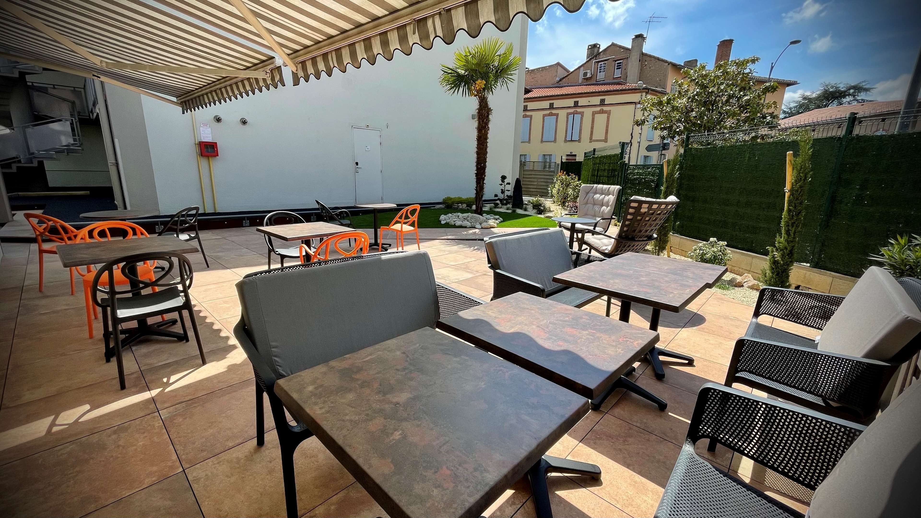 En udendørs terrasse med borde og stole, et palmetræ og en bygning i baggrunden.