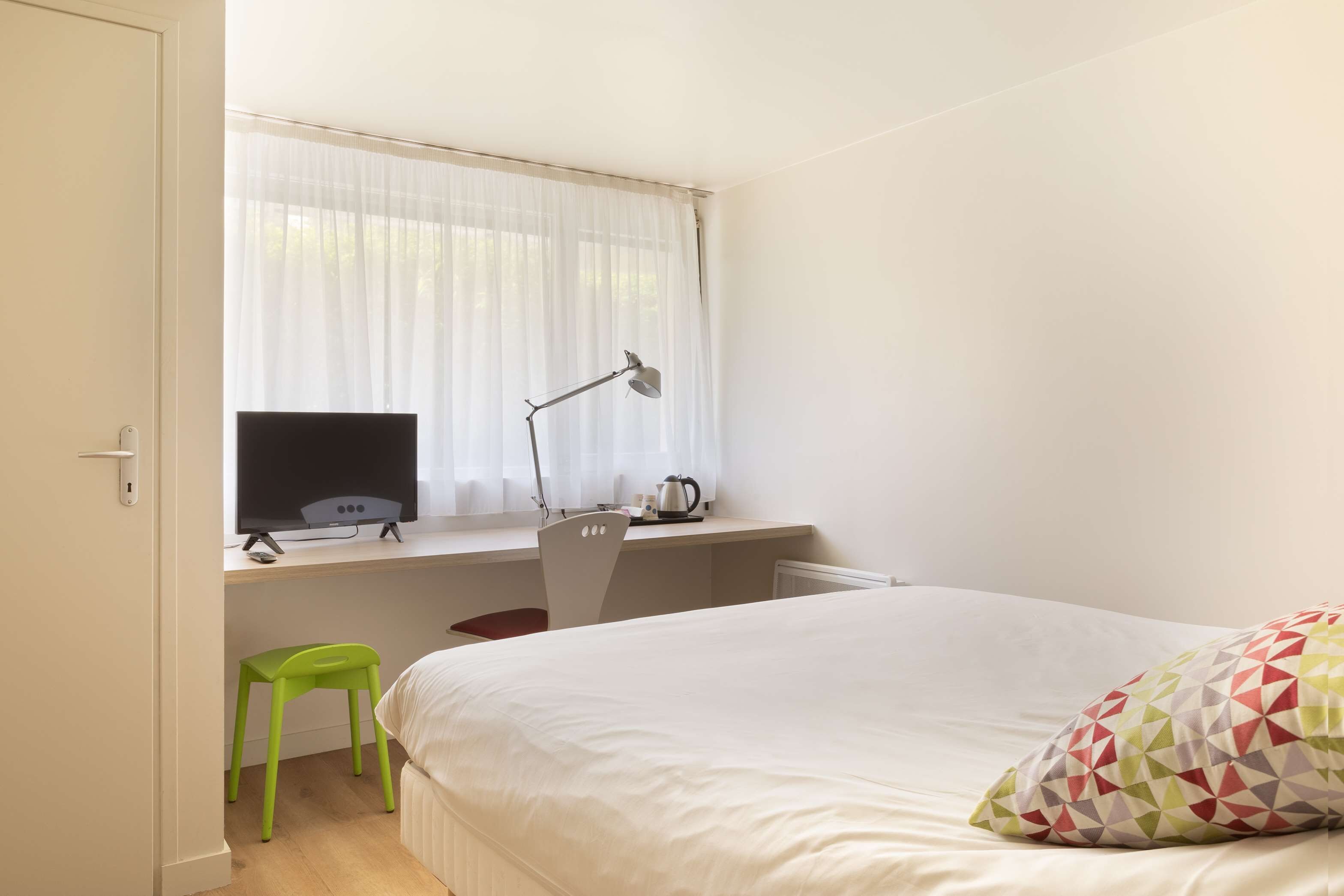 A bedroom with a bed, a desk with a computer, a chair, a window with curtains, and a green stool.