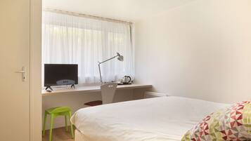 A bedroom with a bed, a desk with a computer, a chair, a window with curtains, and a green stool.