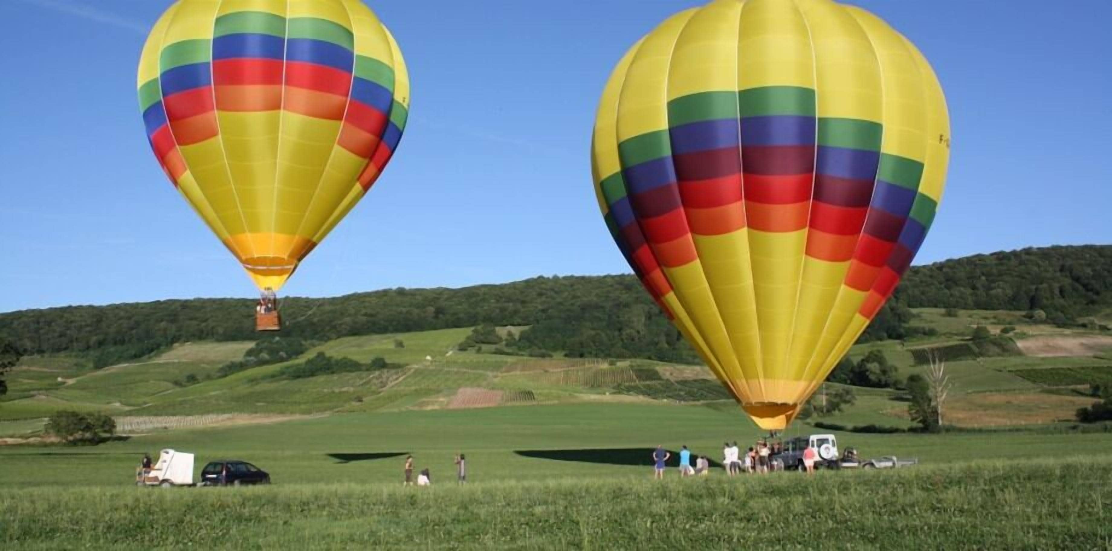 Dos globos aerostáticos sobre un campo, con un grupo de personas y vehículos.
