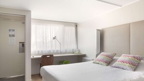 A hotel room with a bed, a desk, a chair, a window with sheer curtains, and a bedside table. - Campanile Dole (Dole)