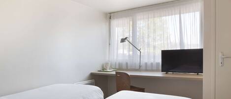 Un dormitorio con dos camas, un escritorio con computadora, una silla y una ventana con cortinas ligeras.