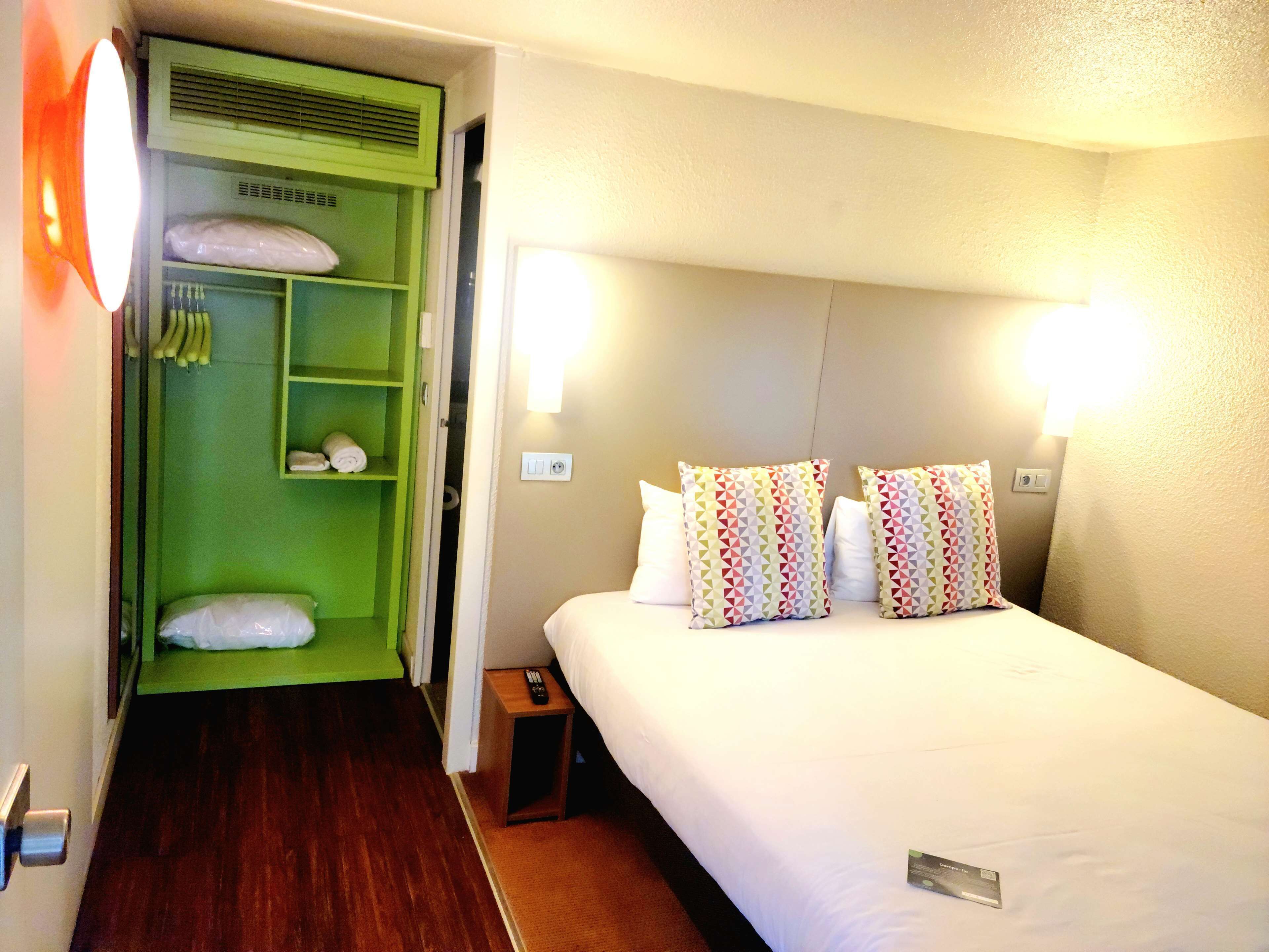 A hotel room with a bed, two pillows, a green wardrobe, and a nightstand with a remote control.