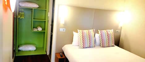 A hotel room with a bed, two pillows, a green wardrobe, and a nightstand with a remote control.