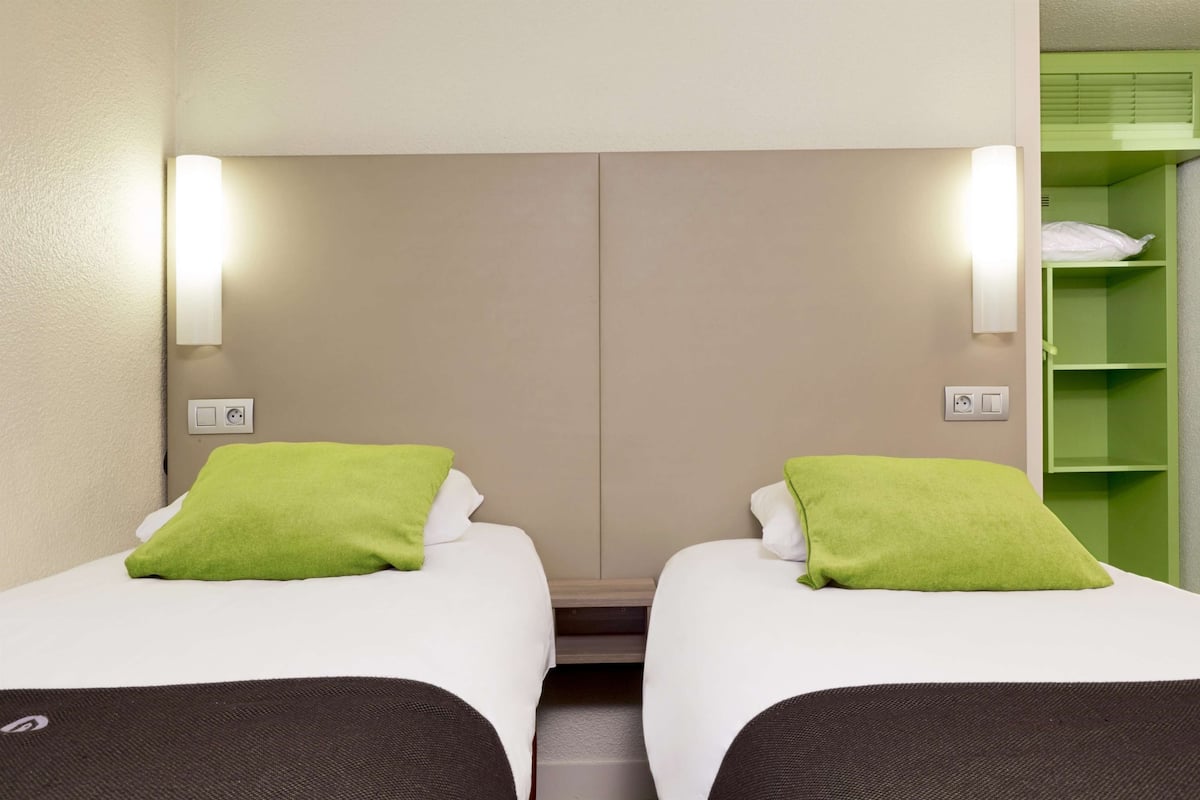 A twin room with two beds, each with a green pillow, a light switch, and a power outlet on the wall.