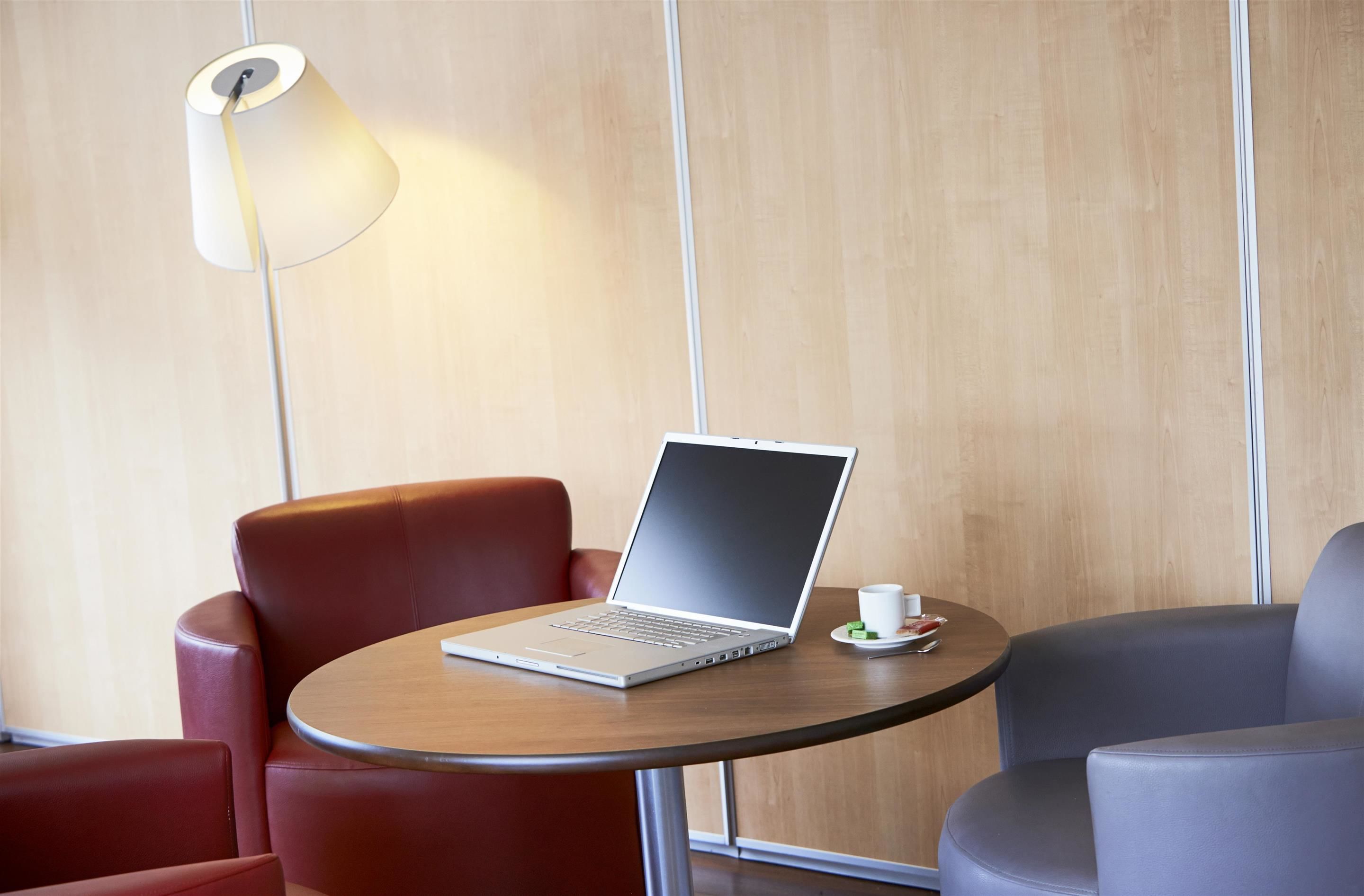 Una habitación moderna con una computadora portátil sobre una mesa redonda, sillas rojas y grises, y una lámpara en la pared.