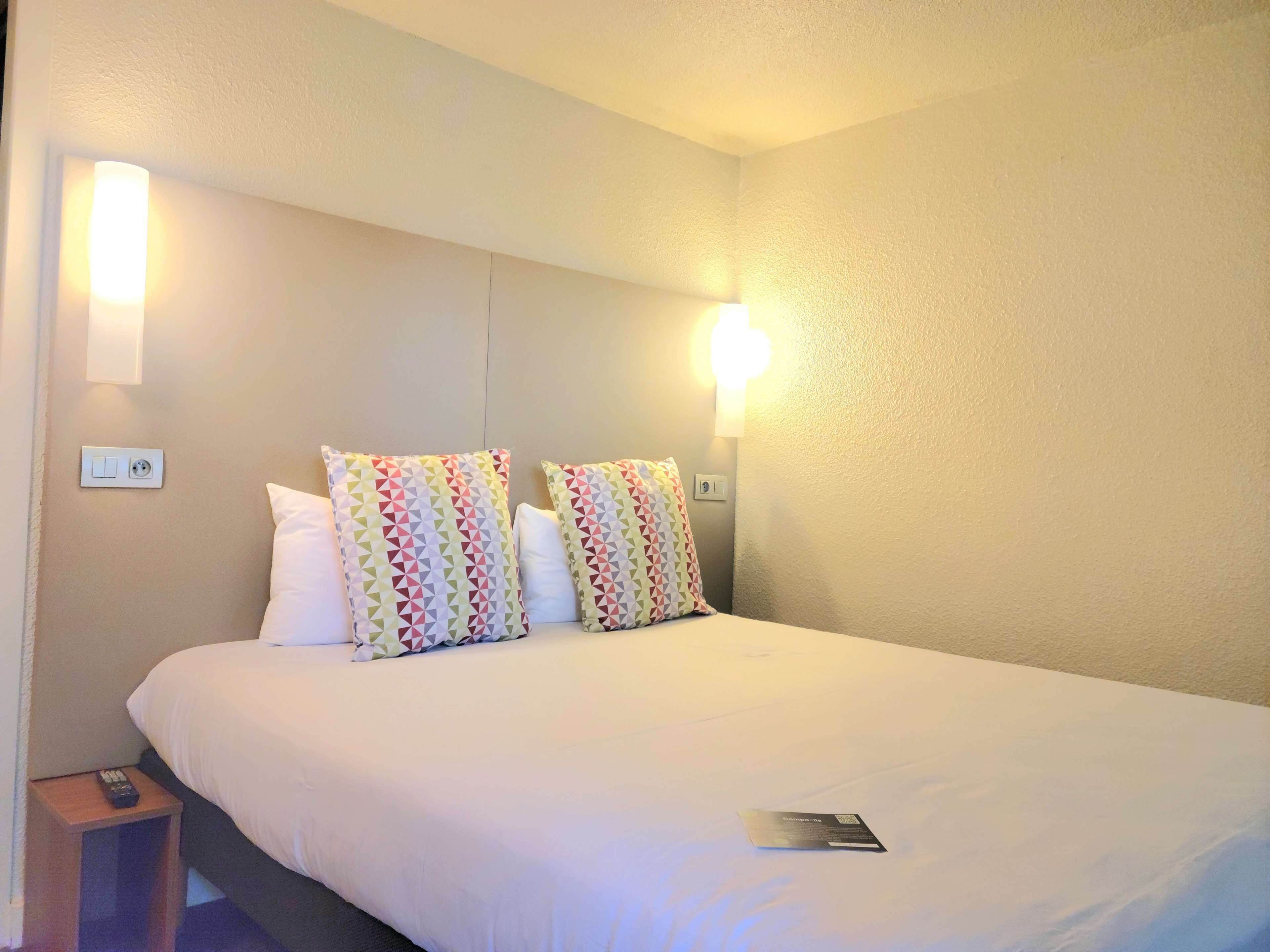 A hotel room with a bed, two pillows, a bedside table with a remote control, and a wall-mounted light fixture.