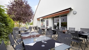 Un espace extérieur aménagé pour les repas, avec des tables et des chaises, un arbre et un bâtiment à la façade blanche.