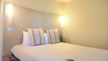 A hotel room with a bed, two pillows, a bedside table with a remote control, and a wall-mounted light fixture.