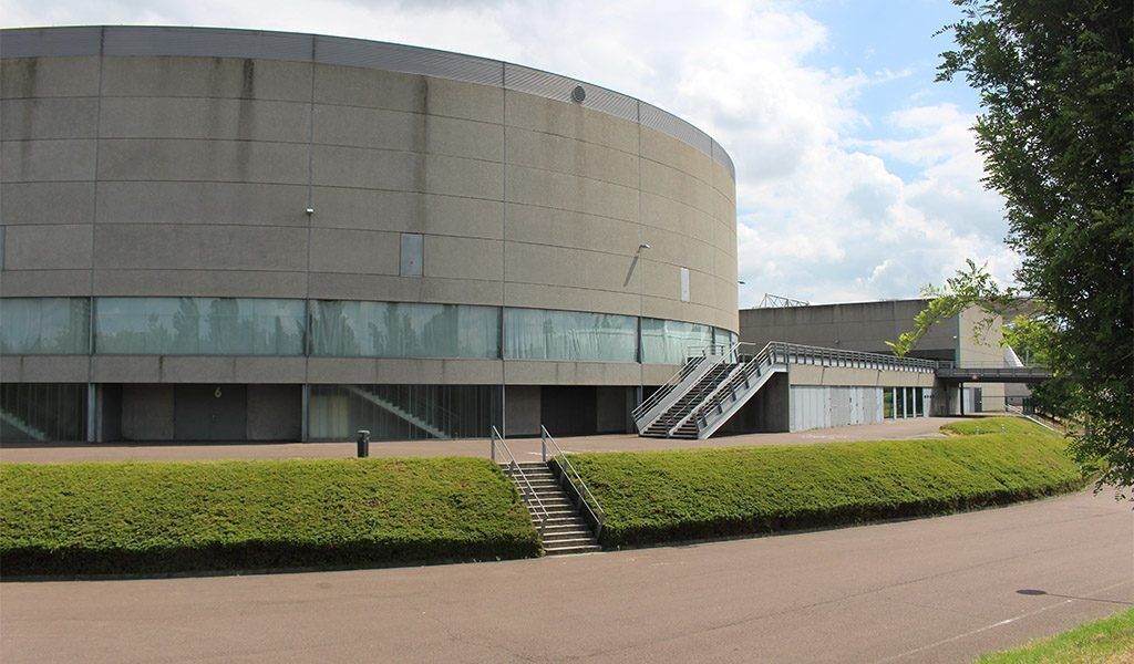 A modern building with a curved facade, a staircase leading to an entrance, and a well-maintained green hedge in front.