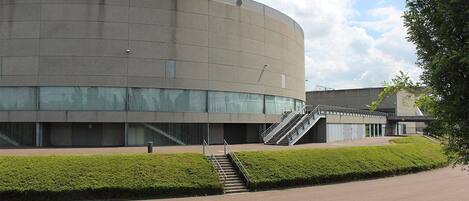 A modern building with a curved facade, a staircase leading to an entrance, and a well-maintained green hedge in front.
