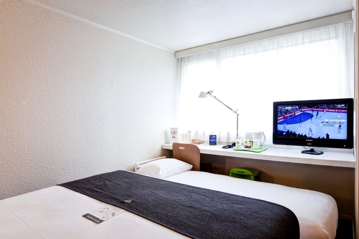 A hotel room with a bed, a desk with a computer monitor, a lamp, and a window with curtains.