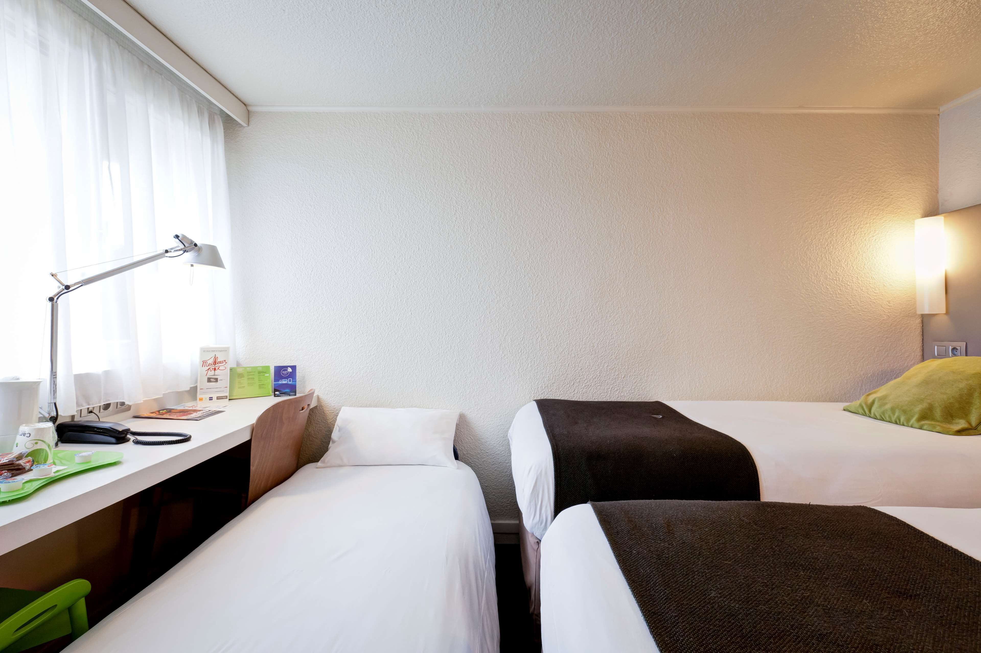 A hotel room with two single beds, a desk with a lamp, a chair, and a window with curtains.