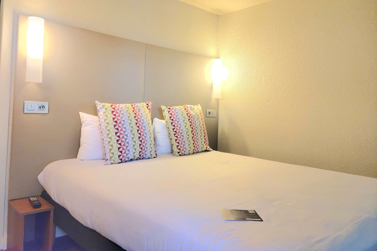 A hotel room with a large bed, two pillows with a geometric pattern, a bedside table with a remote control, and wall-mounted lights.