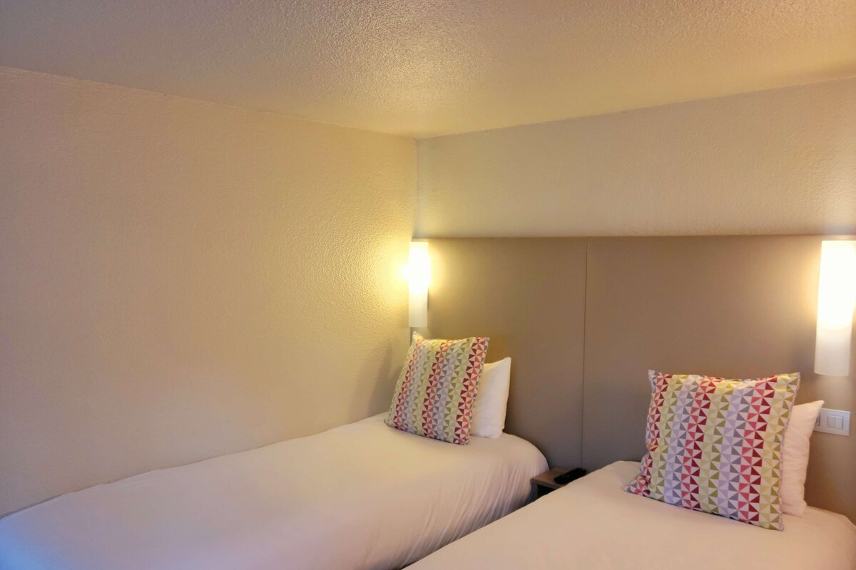 Two single beds with matching headboards and pillows, a bedside table with a remote control, and a wall-mounted light fixture.