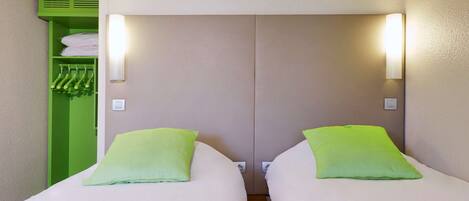 A hotel room with two beds, each with a green pillow and a brown headboard. There is a green storage unit with hanging items and a light switch on the wall.