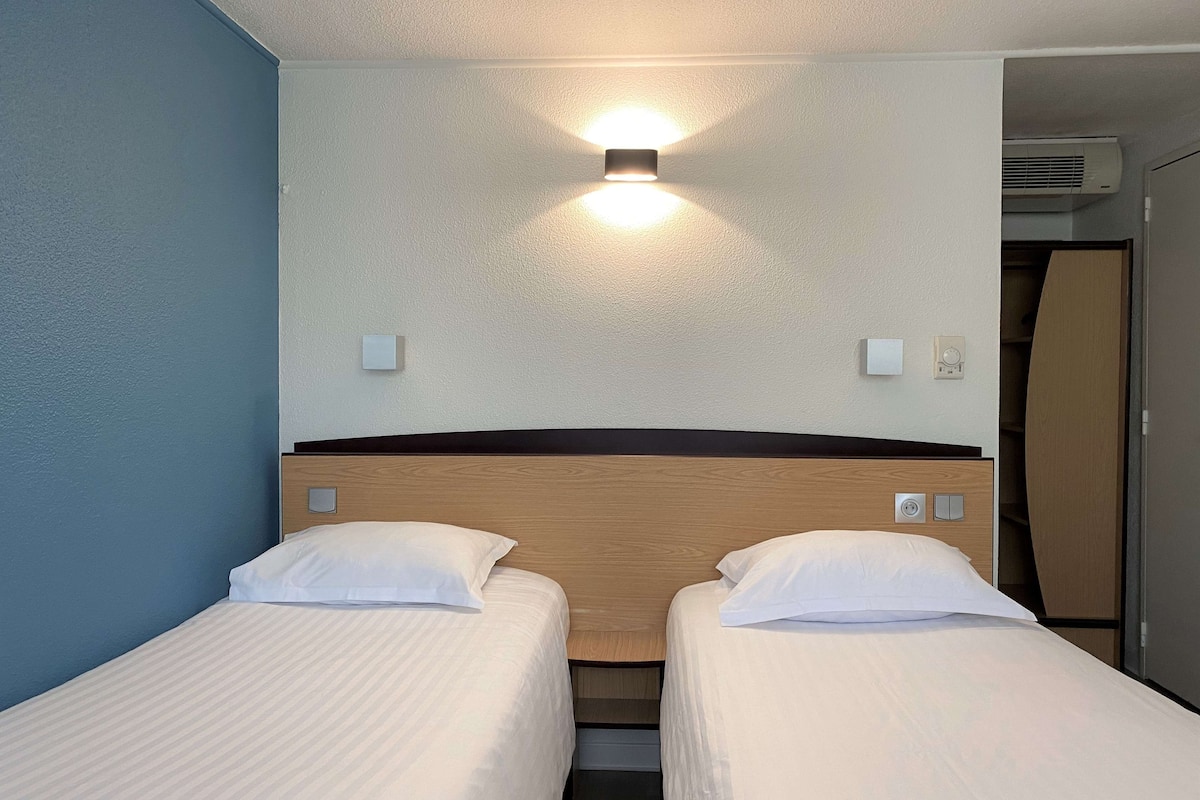 Una habitación de hotel con dos camas individuales, un cabecero de madera, una lámpara de pared y un equipo de aire acondicionado integrado.