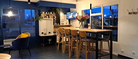 A modern dining area with a wooden table and chairs, a TV mounted on the wall, and a large window offering a view of the outside.