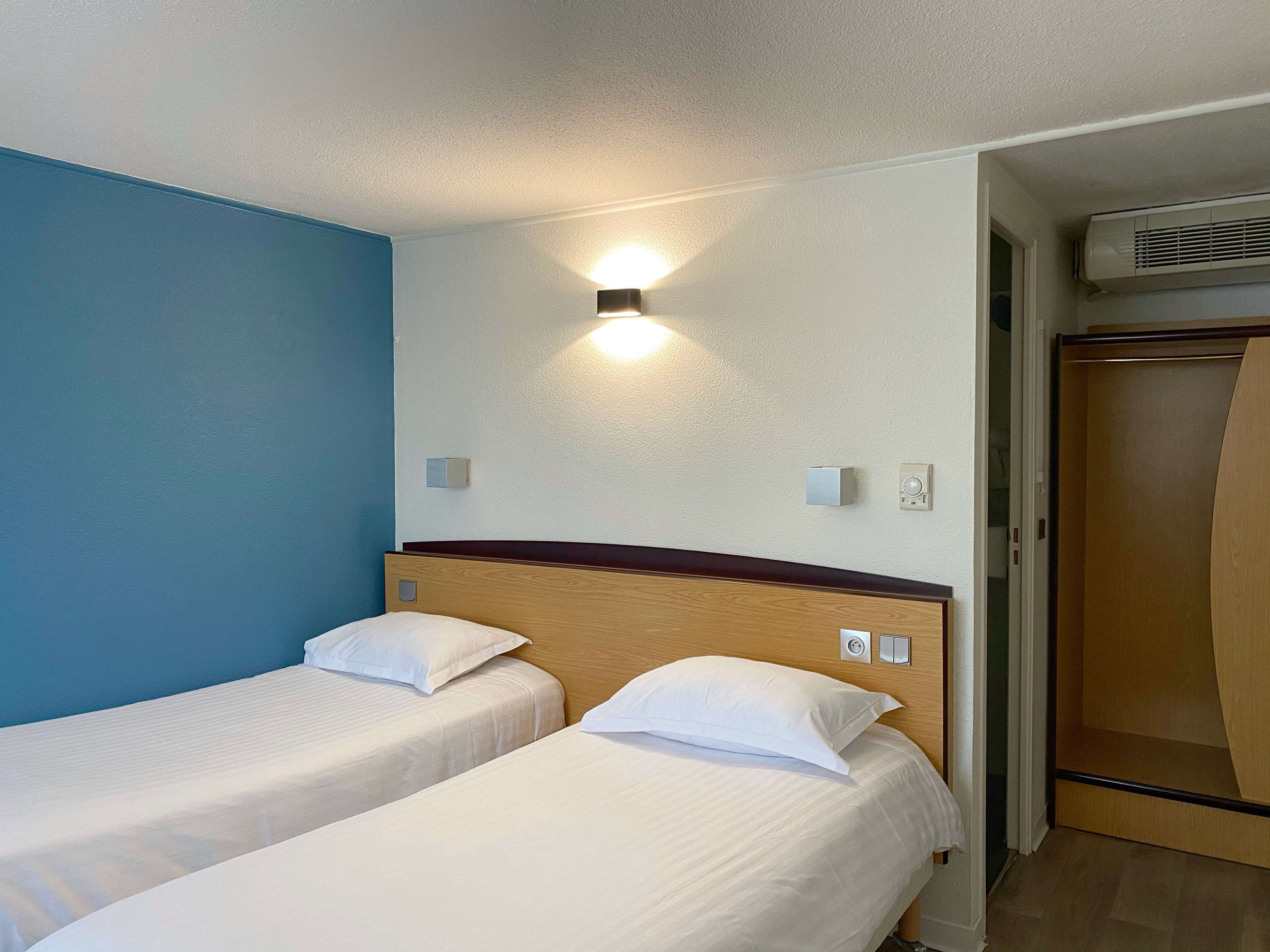 Habitación de hotel con dos camas individuales, una lámpara fijada a la pared, un equipo de aire acondicionado y un armario de madera.